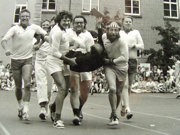 Eine Gruppe in Sportkleidung trägt lachend eine Person über den Schulhof; dahinter sitzen viele Zuschauerinnen und Zuschauer.