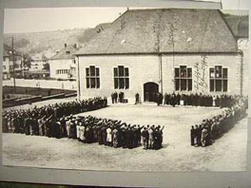 Historische Versammlung vor der Turnhalle; Menschen stehen in Reihen, viele mit erhobenem Arm.
