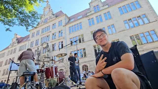 Ein Schüler zeigt neben dem Schlagzeug ein Peace-Zeichen, hinter ihm steht das Schulgebäude.